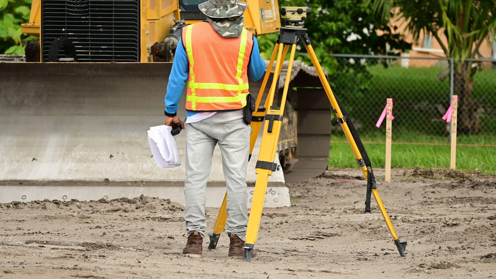 A surveyor with a levelling instrument on a cleared site, marker stakes in the ground.