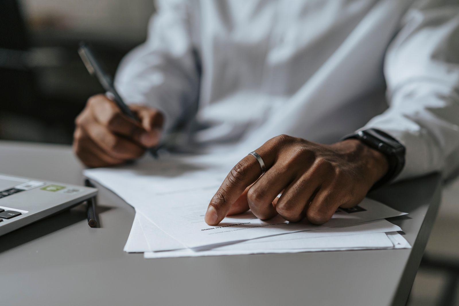 A person signing paperwork at a desk beside a laptop.